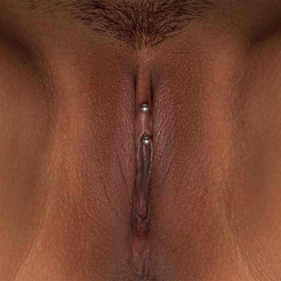 Vulva viewed from underneath. The model has dark skin tone. Labia minora are visible and the clitoris has a piercing. Labia majora skin texture has some bumps. There is no pubic hair on the vulva.